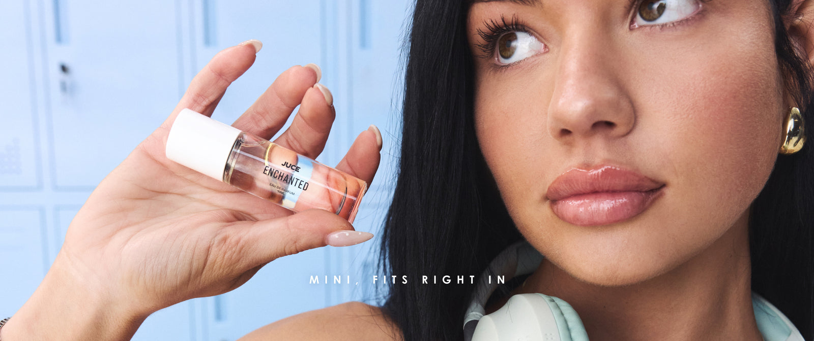 Mini, fits right in - a hand holds a 10ml JUCE Enchanted eau de parfum beside her face, in front of powder-blue lockers.
